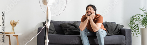 happy bearded man sitting on couch near blurred electric fan, banner photo