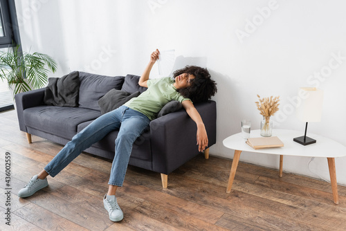 curly african american woman waving with hand fan while lying on grey couch and suffering from heat