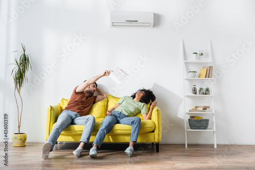 man fixing remote controller near african american girlfriend waving newspaper while suffering from heat