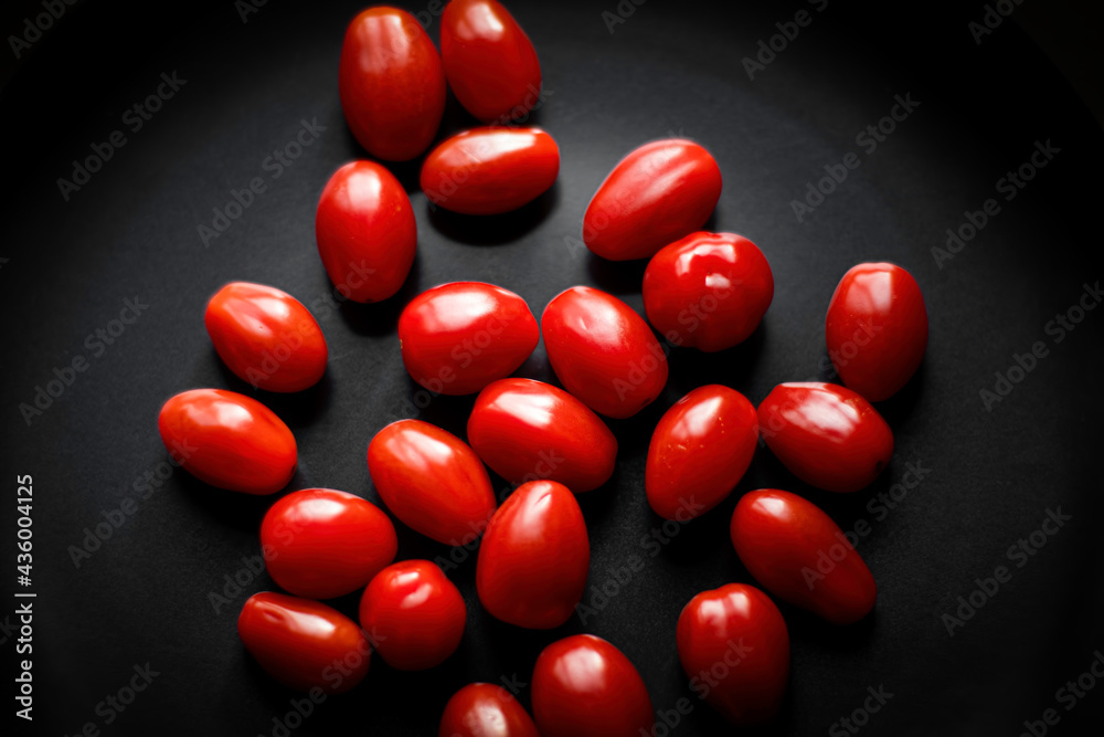 Small oval tomatoes on black ceramic plate. Red vegetables isolated on dark background. Organic garden harvest. Selective focus on the objects, blurred background.