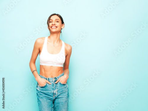 Beautiful smiling woman dressed in white jersey top shirt and jeans. Sexy carefree cheerful model enjoying her morning. Adorable and positive female posing near blue wall in studio