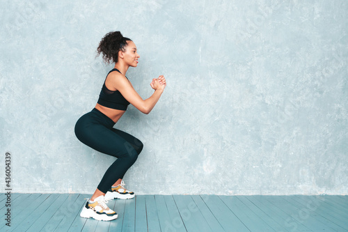 Fitness smiling black woman in sports clothing with afro curls hairstyle.She doing squats. Young beautiful model with perfect tanned body.Female in studio near gray wall. Happy and cheerful