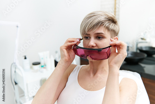 Attractive blonde woman sitting on medical chair in cosmetology room in spa salon and putting on protective UV goggles