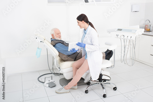 smiling dentist showing tablet to senior patient in dental clinic.