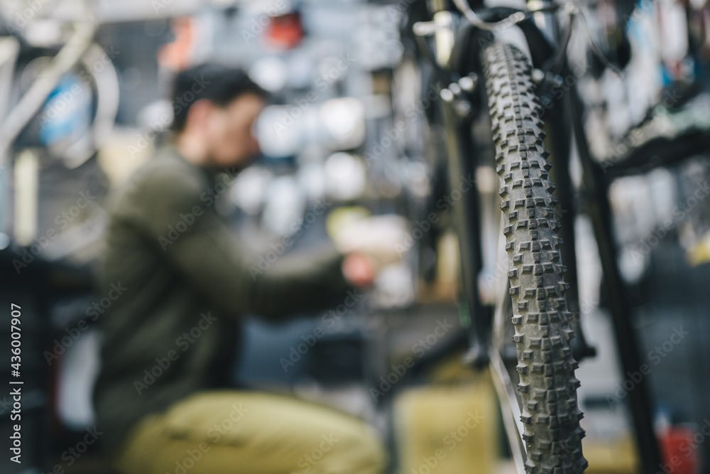 Fototapeta premium Mechanic repairs bicycle in workshop wearing covid 19 mask. Worker of bicycle store fixes bike during coronavirus quarantine in protective mask. Repairman maintenance cycle. Sport shop concept