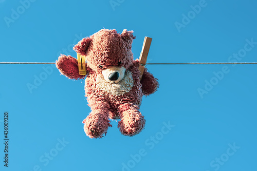 Funny brown teddy bear hanging on a clothesline with wooden clothespin on blue sky background.
