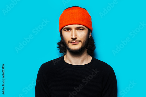 Studio portrait of young guy on blue background, wearing orange hat and black sweater. © Lalandrew
