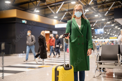 Young woman in protective mask in green coat holds passport with ticket and yellow suitcase airport. Concept of traveling by plane during covid virus