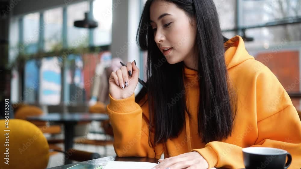 The pensive woman in an orange hoodie writing something in a notebook from the phone while sitting in a cafe The pensive woman in an orange hoodie writing something in a notebook from the phone while sitting in a cafe
