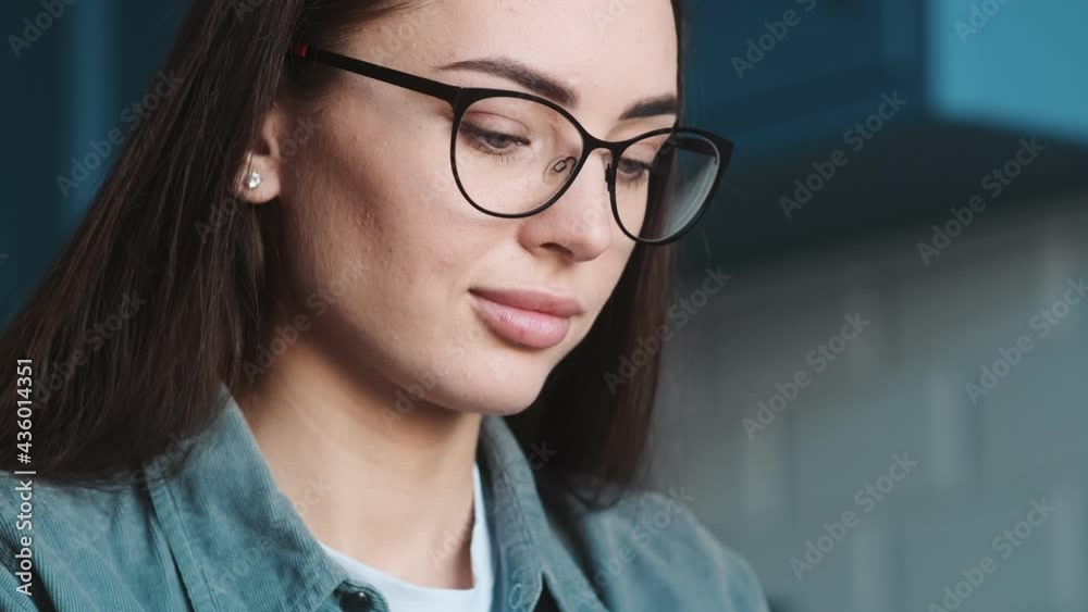 The close-up view of the concentrated woman with glasses reading something and looking down while sitting at the table in the kitchen The close-up view of the concentrated woman with glasses reading something and looking down while sitting at the table in the kitchen
