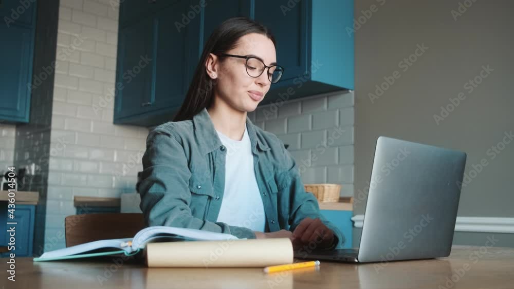The smiling woman in glasses getting ready for a video call conversation on the laptop and waving hand to his camera in a kitchen The smiling woman in glasses getting ready for a video call conversation on the laptop and waving hand to his camera in a kitchen