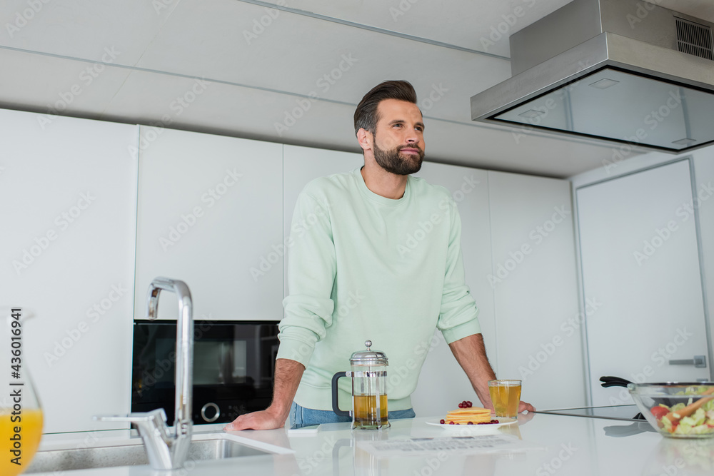 positive man looking away near green tea and delicious pancakes in kitchen positive man looking away near green tea and delicious pancakes in kitchen
