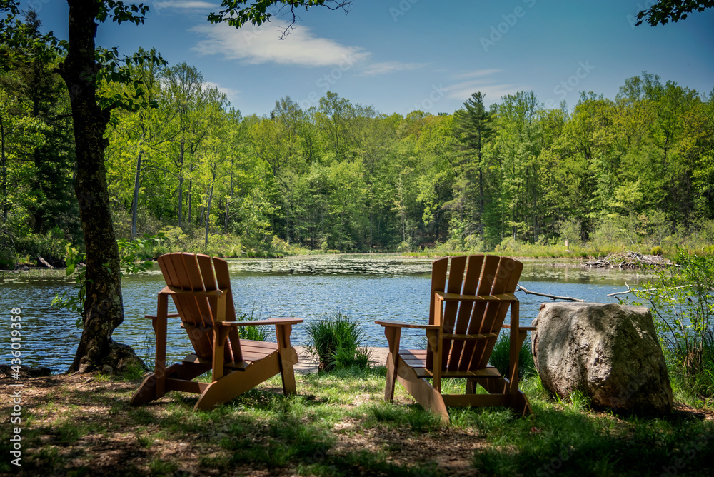 Relaxing inviting scene on summer day with two chairs facing a pond with forest in background and blue sky Relaxing inviting scene on summer day with two chairs facing a pond with forest in background and blue sky