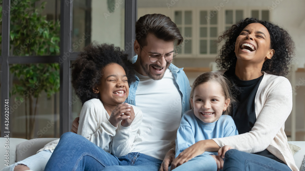 Overjoyed young multiracial family with two small daughters relax on sofa at home laughing joking. Happy multiethnic mom and dad rest together with small diverse children. Adoption concept. Overjoyed young multiracial family with two small daughters relax on sofa at home laughing joking. Happy multiethnic mom and dad rest together with small diverse children. Adoption concept.
