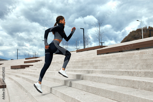 Young woman runner athlete in dark skinny suit running down the city stairs, jogging and jogging at city training workout