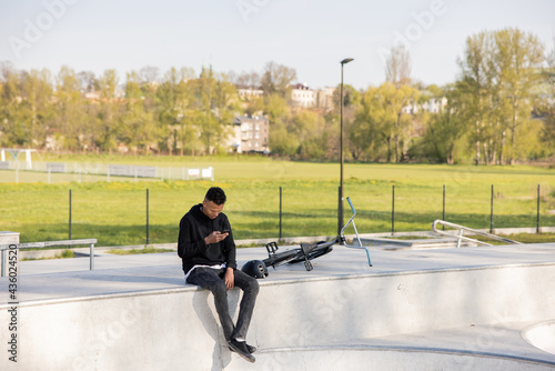 Boy sits in helmet with bmx bike in park on ramp, at skatepark with phone in hand and texts friends he hangs out with to do tricks photo