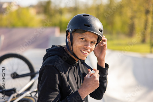 A young boy sits on a ramp at a skatepark with his bike lying wheel up behind him. The guy has finished riding and removes a black helmet from his head, unzips it from under his neck