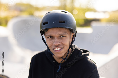 A young wearing a helmet stares into the camera with dark brown eyes with white, straight teeth on top. In the background you can see the ramp and the greenery of the park surrounding the ramp © ABCreative