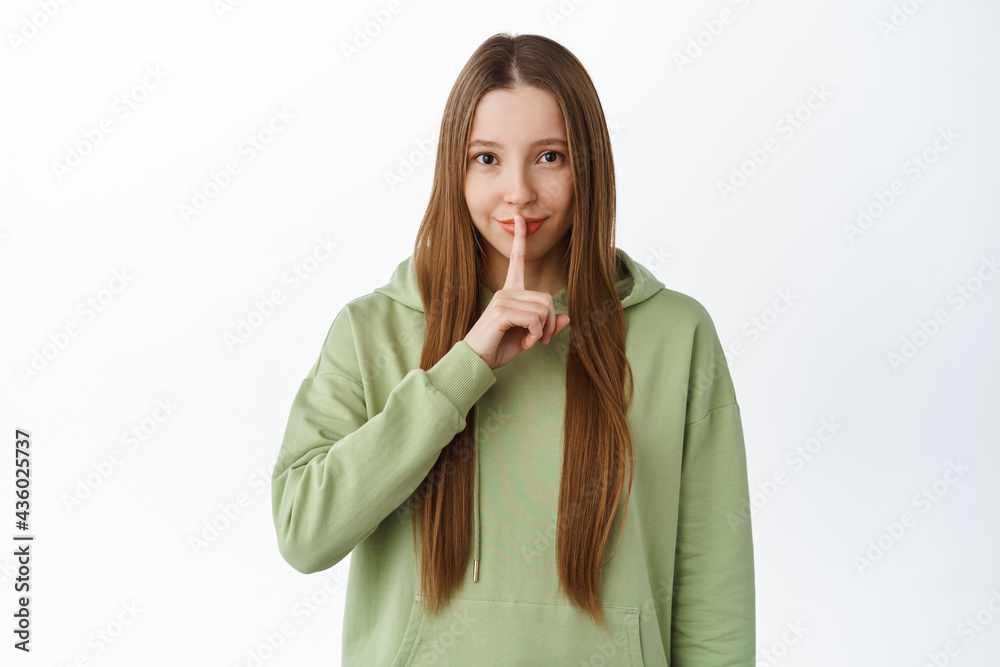 Smiling young woman make hush, shhh gesture, asking to keep quiet, voice down, make taboo shush gesture, standing over white background in green hoodie Smiling young woman make hush, shhh gesture, asking to keep quiet, voice down, make taboo shush gesture, standing over white background in green hoodie