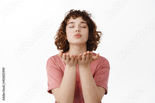 Romantic young woman close eyes  sending air kiss  blowing mwah kissing in open hands near lips  standing in t-shirt against white background