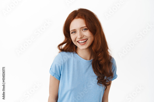 Portrait of attractive redhead woman with curly hair, smiling with white teeth, gazing happy at camera, wearing blue t-shirt against white background