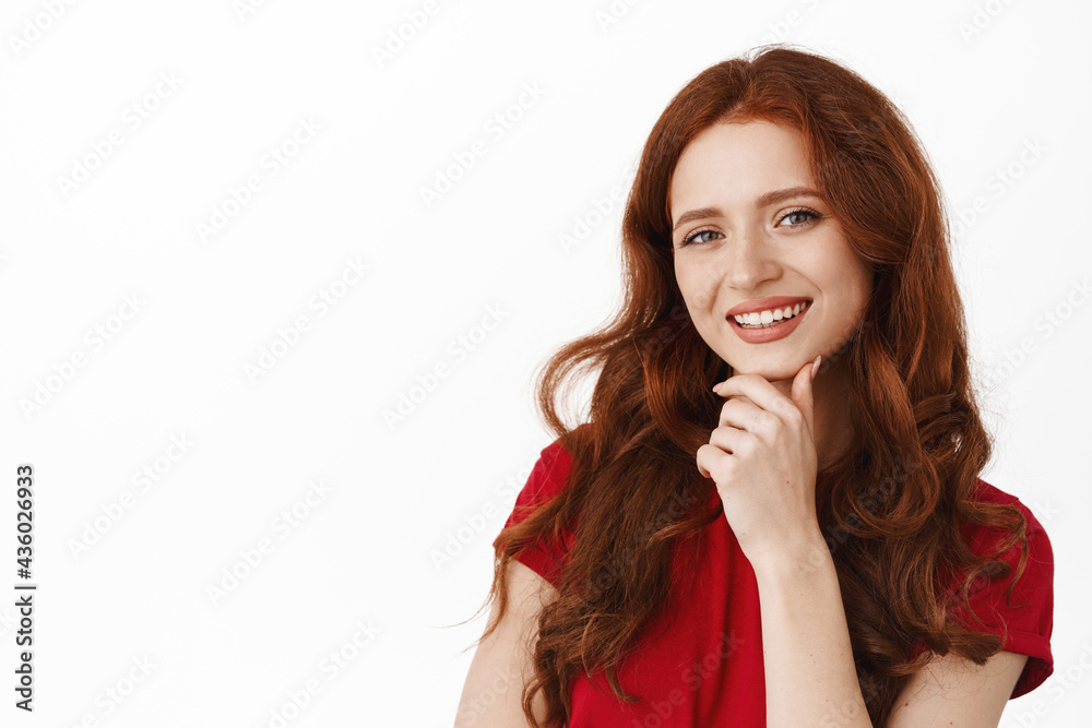 Portrait of successful smiling redhead girl with long curly hair, touch chin, looking pleased and satisfied at something interesting, standing thoughtful against white background Portrait of successful smiling redhead girl with long curly hair, touch chin, looking pleased and satisfied at something interesting, standing thoughtful against white background