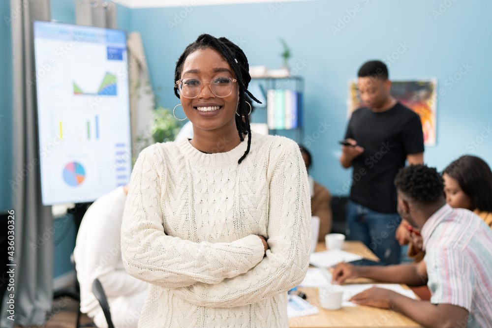Portrait of a smiling dark-skinned girl wearing a warm sweater with her arms crossed over chest, woman wears glasses and gold earrings, her hair is braided, a business meeting in office in background - obrazy, fototapety, plakaty 