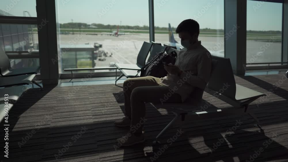 Masked man sits at airport on social distance seat by window before boarding plane and use his phone out of boredom. Flight delay. Cancellation of flights due to quarantine and lockdown covid 19 Masked man sits at airport on social distance seat by window before boarding plane and use his phone out of boredom. Flight delay. Cancellation of flights due to quarantine and lockdown covid 19