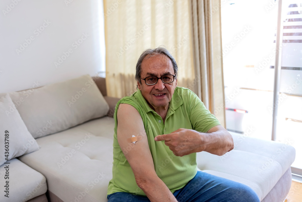 Portrait of a proud cheerful senior man who had just been vaccinated sitting on sofa at home. Senior man showing vaccinated arm. Coronavirus vaccination concept of man received the COVID-19 vaccine. Portrait of a proud cheerful senior man who had just been vaccinated sitting on sofa at home. Senior man showing vaccinated arm. Coronavirus vaccination concept of man received the COVID-19 vaccine.