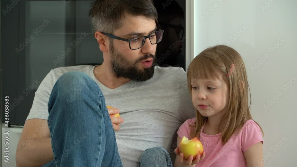 Happy family, smiling father and little daughter sit on floor in kitchen at home and eat ripe apples. Child bites fruit. Food concept Happy family, smiling father and little daughter sit on floor in kitchen at home and eat ripe apples. Child bites fruit. Food concept