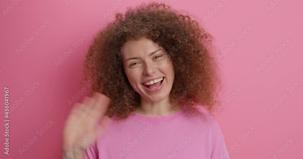 Overjoyed curly haired young European woman waves palm in hello gesture greets best friend blows air kiss smiles gladfully being in good mood dressed in casual clothes isolated over pink background. Overjoyed curly haired young European woman waves palm in hello gesture greets best friend blows air kiss smiles gladfully being in good mood dressed in casual clothes isolated over pink background.