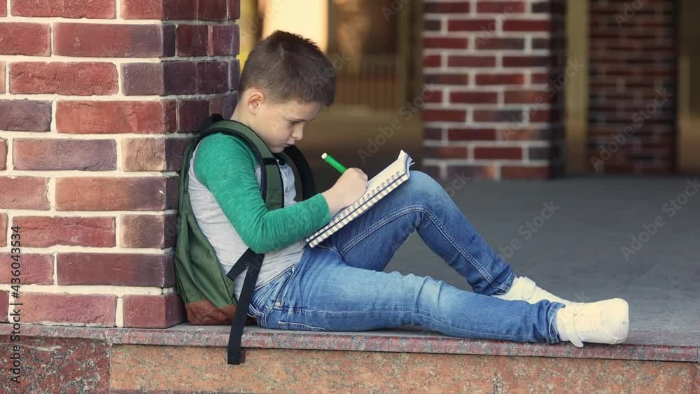 School boy in casual clothes sitting in the schoolyard against a brick wall doing homework. The child drawing in a notebook with a marker pen and waiting for parents after lessons. School boy in casual clothes sitting in the schoolyard against a brick wall doing homework. The child drawing in a notebook with a marker pen and waiting for parents after lessons.