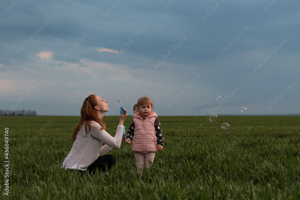 Mom plays with her daughter in the green field. A little girl watches her mother blow bubbles Mom plays with her daughter in the green field. A little girl watches her mother blow bubbles