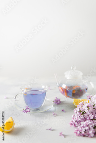 Cup of purple tea with lemon, teapot, bouquet of blooming lilac on light background. Spring tea drinking. Greeting card, invitation design. Cafe menu, poster. Lilac tea recipe. Side view, copy space