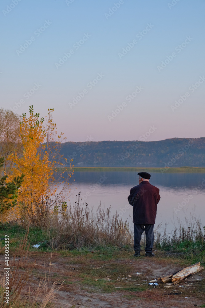 Summer, river, sunset, landscape. An elderly man stands in the foreground and looks to the other side. Loneliness.