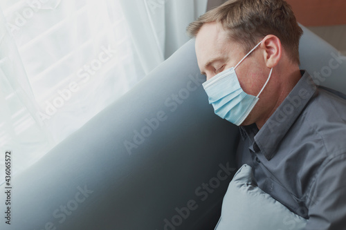 Young attractive Caucasian men relaxing wearing protective face mask having a nap on couch in the afternoon. Happy employee man resting with eyes closed on the sofa during state quarantine