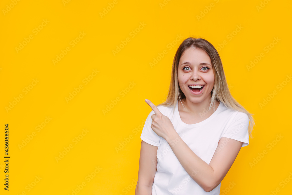 A young caucasian smiling impressed blonde woman in a white t-shirt indicate copy empty blank space for text or design with her finger presenting product isolated on a bright color yellow background A young caucasian smiling impressed blonde woman in a white t-shirt indicate copy empty blank space for text or design with her finger presenting product isolated on a bright color yellow background