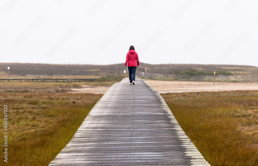 Lonely woman walking on a wooden walkway through a tidal marsh on a misty autumn day. Concept of loneliness.