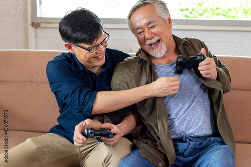 Son and dad fight for gamepad in cozy home, smiling and laughing together. Senior Asian man challenge with Young Asian man gamer. Father's day and happy family concept.