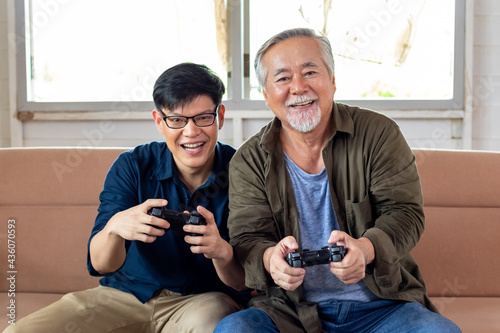 Son and dad enjoy playing video game together sitting on sofa in cozy home, smiling and laughing together. Senior Asian man having fun time with Young Asian man. Father's day and happy family concept.