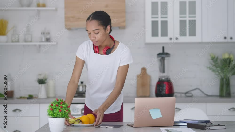 Wide shot of young slim woman bringing plate with fruits on kitchen table and sitting down to laptop. Serious African American freelancer start working online in home office indoors in the morning Wide shot of young slim woman bringing plate with fruits on kitchen table and sitting down to laptop. Serious African American freelancer start working online in home office indoors in the morning