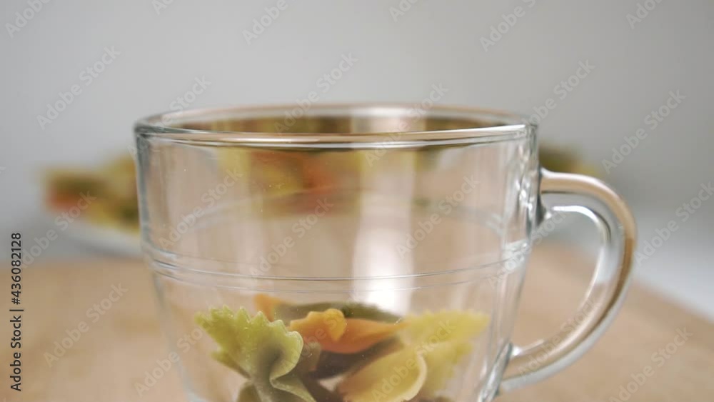 Farfalle multicolored uncooked pasta in the shape of a butterfly falls into a glass cup. Slow motion. Close up Farfalle multicolored uncooked pasta in the shape of a butterfly falls into a glass cup. Slow motion. Close up