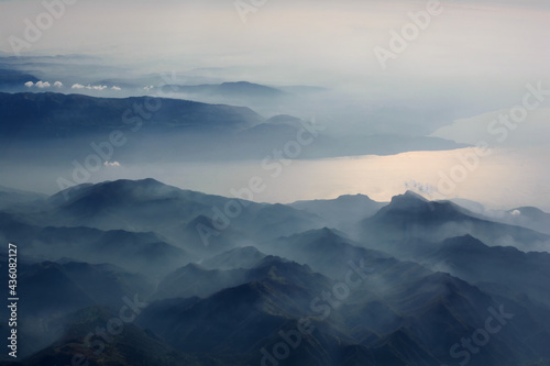 Aerial view of misty mountains, lake and clouds above the mountain peaks, opposite the sunlight, blue tinted © DLVV
