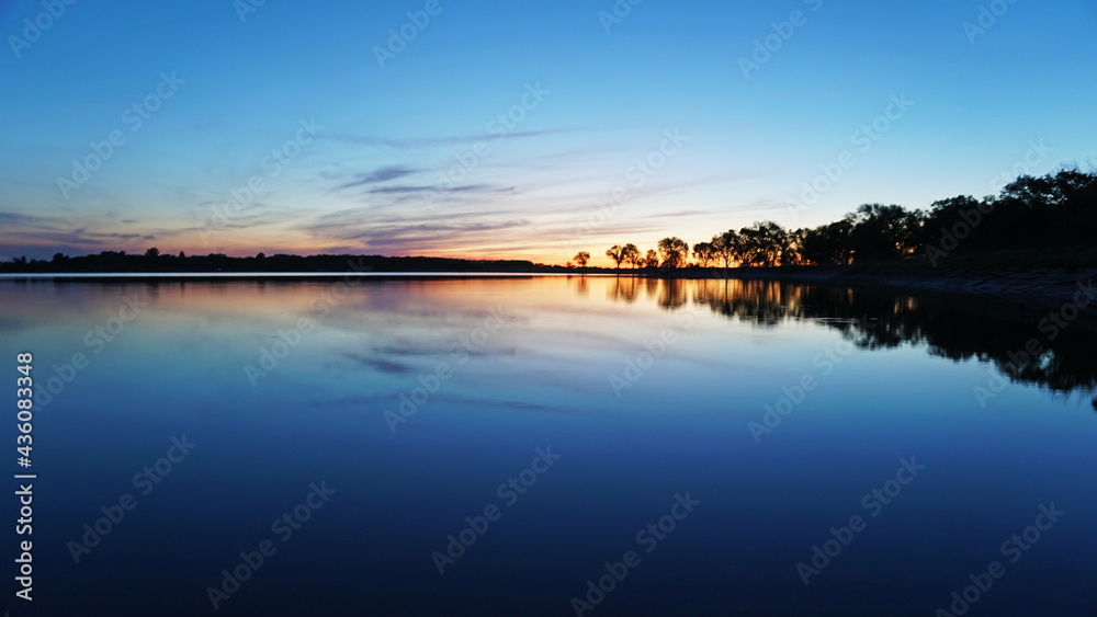 Sunset overlooking the lake, trees, clouds. The sky and bushes are reflected in the water. Landscape at dusk. Nightfall. Water like a mirror.