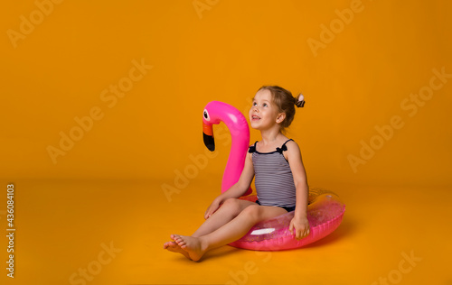 little blonde girl in a striped swimsuit sits in a pink inflatable circle with a flamingo and looks away on a yellow background