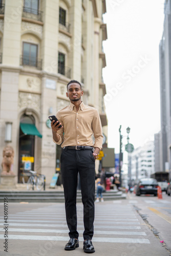 Full length shot of African businessman outdoors in city smiling and using mobile phone