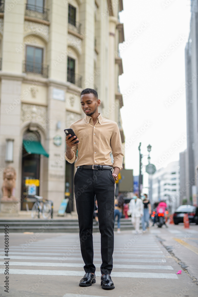 Full body shot of African businessman outdoors in city smiling and using mobile phone
