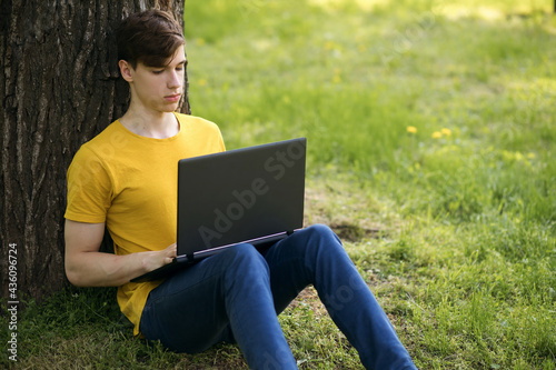 A young student guy sits on the lawn and holds a laptop in his hands. Rest after school or work. Remote work of a young man using a laptop outdoors. Free space for text