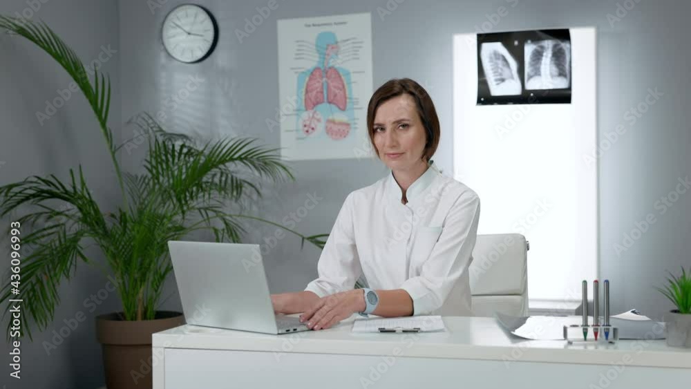 Female Doctor Sitting At The Table And Using Laptop. Young Woman In White Coat Sitting In The Office. Female Doctor Sitting At The Table And Using Laptop. Young Woman In White Coat Sitting In The Office.