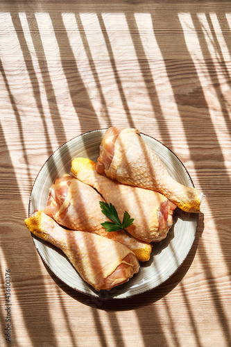 Vertical composition of the top view of a plate with raw chicken legs on a wooden table next to a window that lets in the rays of the sun between lights and shadows